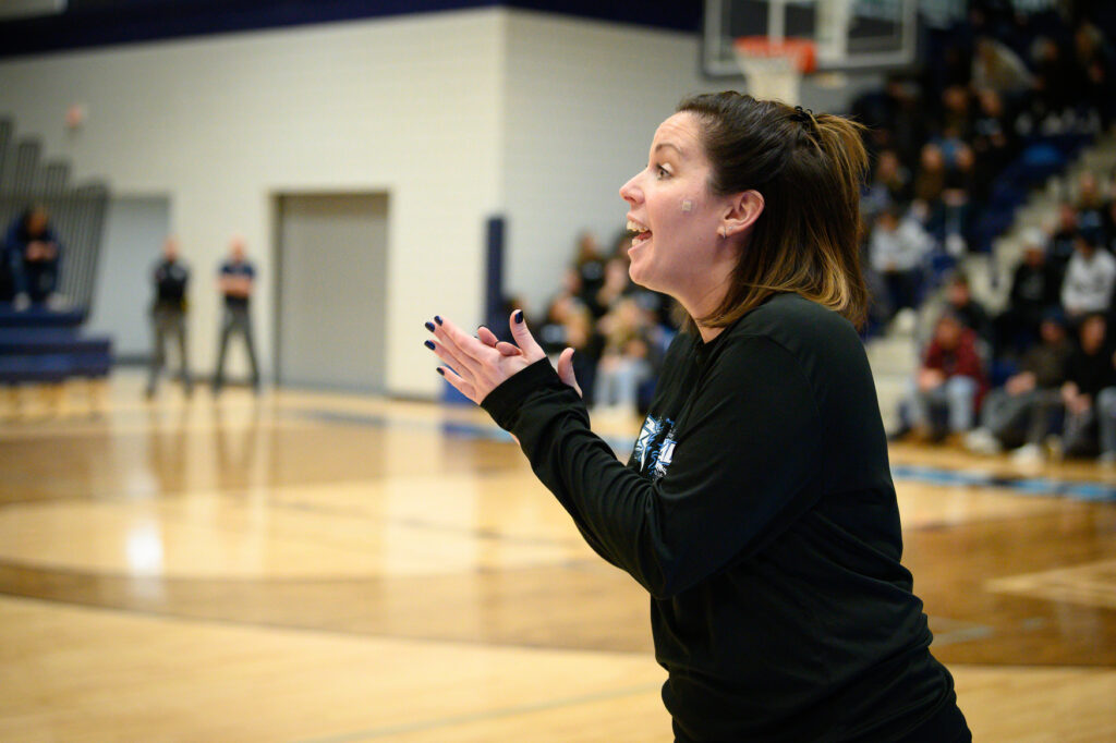 A woman in a black long-sleeve shirt claps and speaks on the sidelines of a basketball court, possibly involved in sports marketing, with spectators visible in the background.