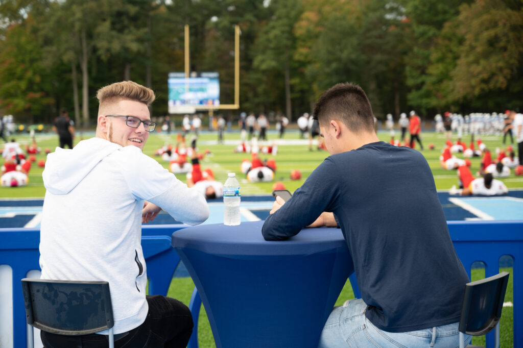Two young men sit at a blue table overlooking a football field where players are stretching; one person looks back and smiles at the camera, perfectly capturing an authentic moment for sports marketing.