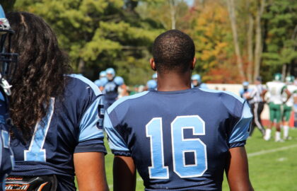 Two American football players in blue jerseys, numbers 51 and 16, stand on the sideline watching a game on a sunny day.