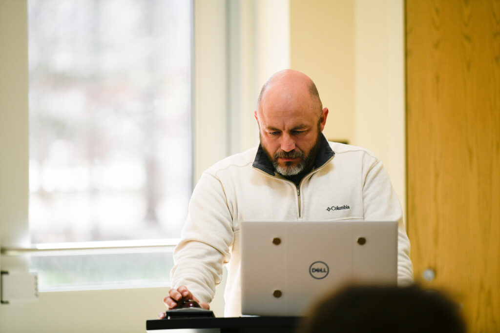 A man with a bald head and beard, wearing a light-colored Columbia fleece, stands at a podium using a Dell laptop in a brightly lit room, preparing for his sports marketing presentation.