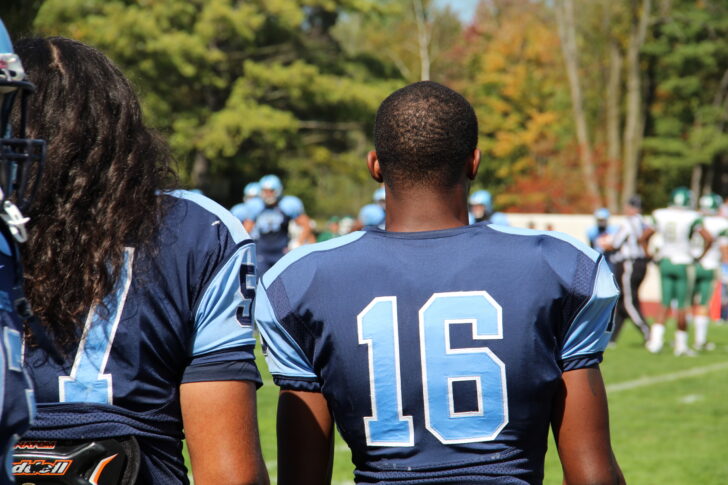 Two American football players in blue jerseys, numbers 51 and 16, stand on the sideline watching a game on a sunny day.