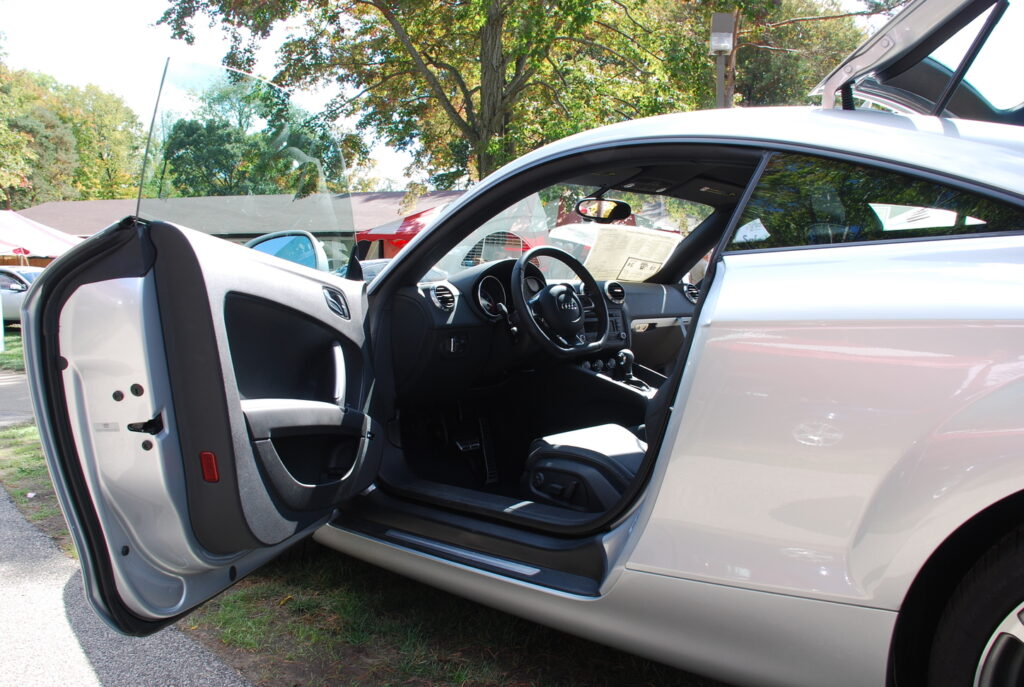 A silver sports car, perfect for automotive marketing, is parked outdoors with its driver-side door open, revealing the sleek black interior and dashboard under a tree on a sunny day.