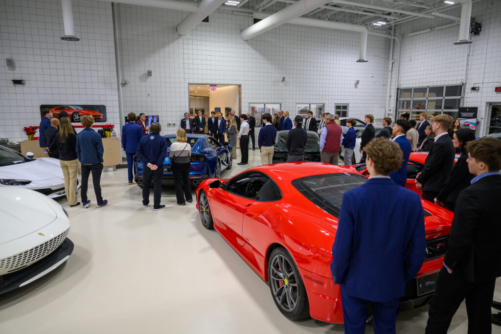 A group of people stands and converses inside a bright showroom with several high-end sports cars, including a red Ferrari, visible in the foreground.