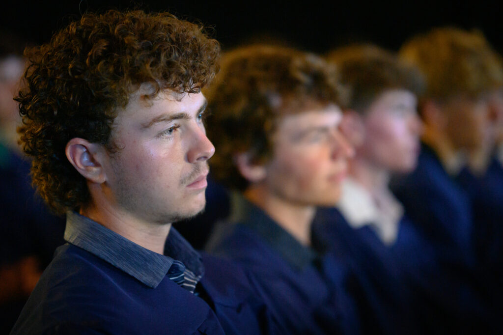 A young man with curly hair sits among a row of people, all wearing dark clothing, attentively facing forward in a dimly lit setting—perhaps gathered for an automotive marketing presentation.