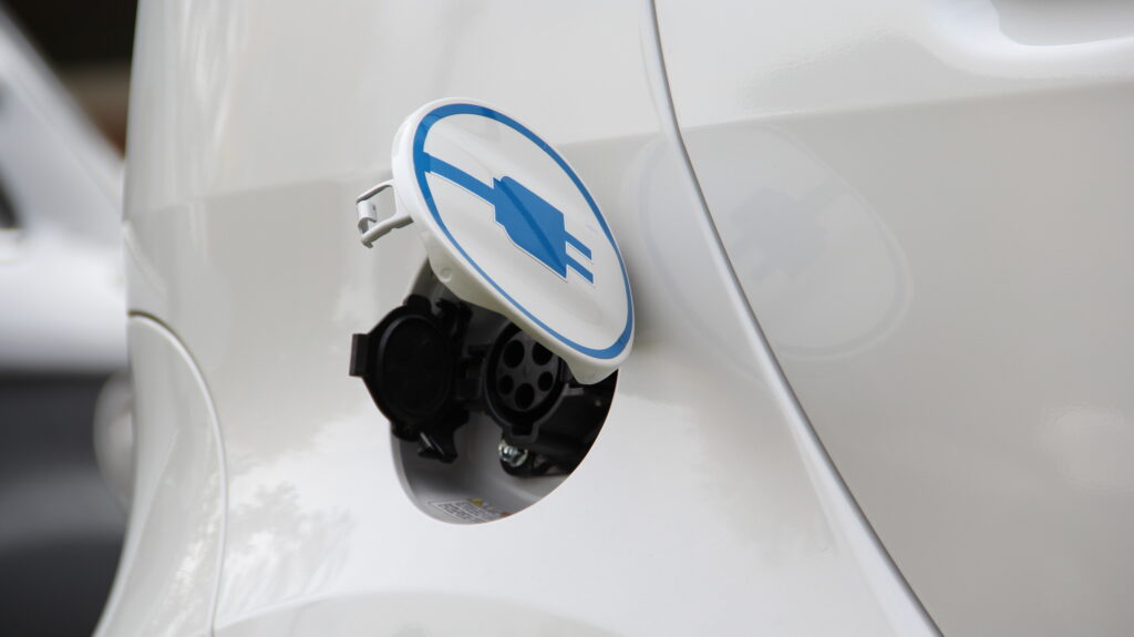 Close-up of an electric vehicle charging port with the cover open, showing the charging socket and a blue electric plug symbol—perfect for automotive marketing campaigns focused on sustainability and innovation.