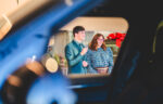 A man and a woman stand indoors, viewed through a car window, smiling in business attire—capturing a moment that embodies professionalism and the essence of automotive marketing.