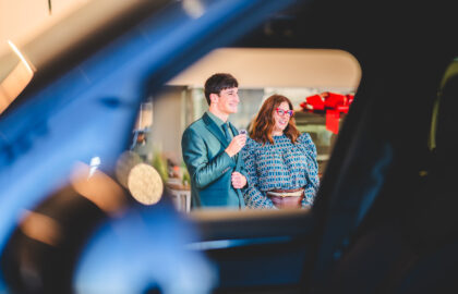 A man and a woman stand indoors, viewed through a car window, smiling in business attire—capturing a moment that embodies professionalism and the essence of automotive marketing.
