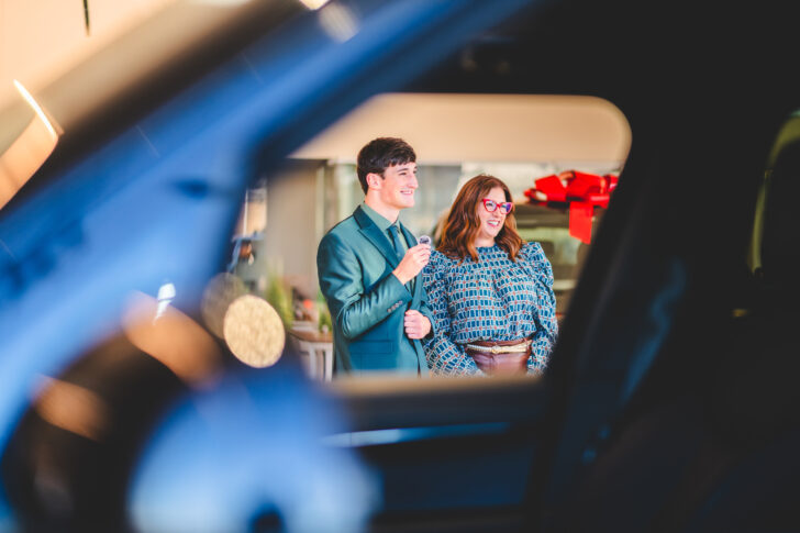 A man and a woman stand indoors, viewed through a car window, smiling in business attire—capturing a moment that embodies professionalism and the essence of automotive marketing.