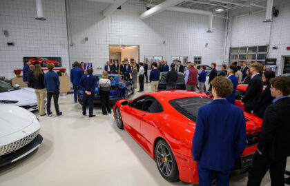 A group of people stands and converses inside a bright showroom with several high-end sports cars, including a red Ferrari, visible in the foreground.