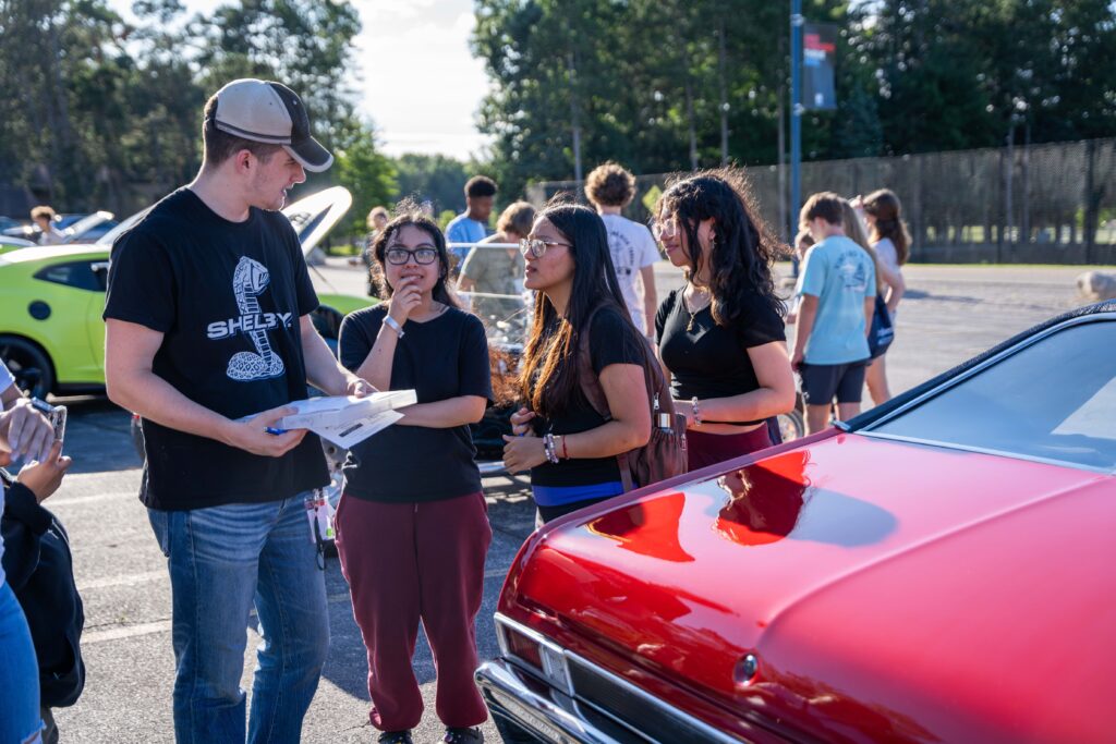 A group of people stand by a red car in a parking lot, with one person holding papers and discussing automotive marketing strategies. Other cars and people are visible in the background.