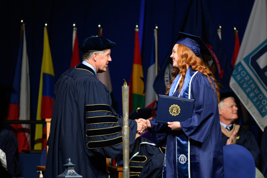 A graduate in cap and gown receives a Bachelor Degree diploma and handshake from a faculty member on stage at a graduation ceremony, with flags in the background.