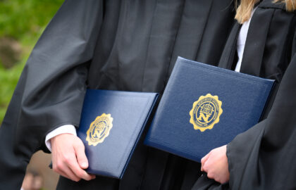 Two graduates in black robes hold blue diploma covers with a gold university seal during an outdoor graduation ceremony.