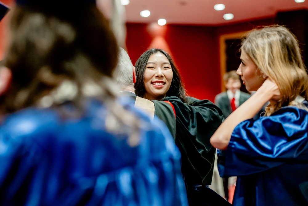 A graduate in a black gown, celebrating their bachelor degree, smiles while hugging someone, surrounded by others in blue gowns at a graduation ceremony.