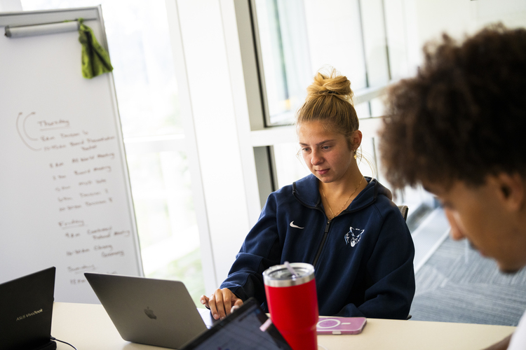 A young woman pursuing her bachelor degree works on a laptop at a table with others; a whiteboard with handwritten notes is visible in the background.