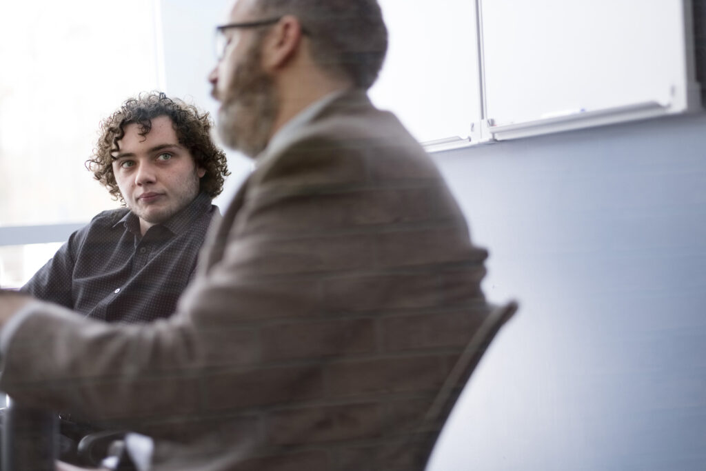 Two men sit at a table in an office, one with curly hair listening attentively as the other, wearing glasses and a jacket, discusses his bachelor degree.