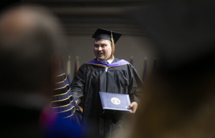 A graduate in cap and gown holds a diploma and smiles while walking across a stage during a graduation ceremony.