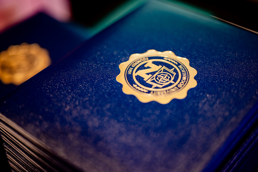 A close-up of a blue bachelor degree certificate folder with a gold embossed seal on the cover, stacked with others in the background.