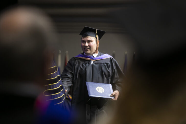 A graduate in cap and gown holds a diploma and smiles while walking across a stage during a graduation ceremony.