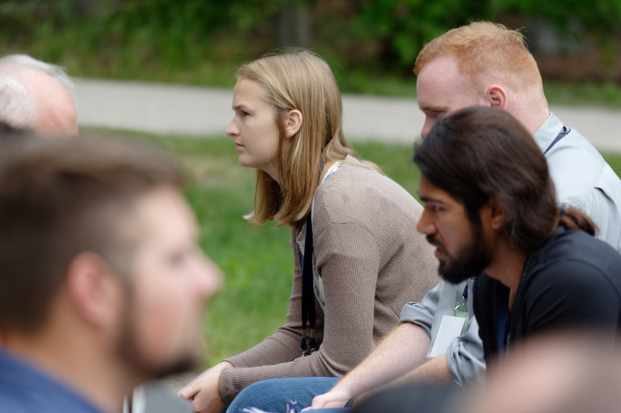 A group of people sitting outdoors, focused and listening, with greenery in the background—engaged in a discussion about online classes for adults.