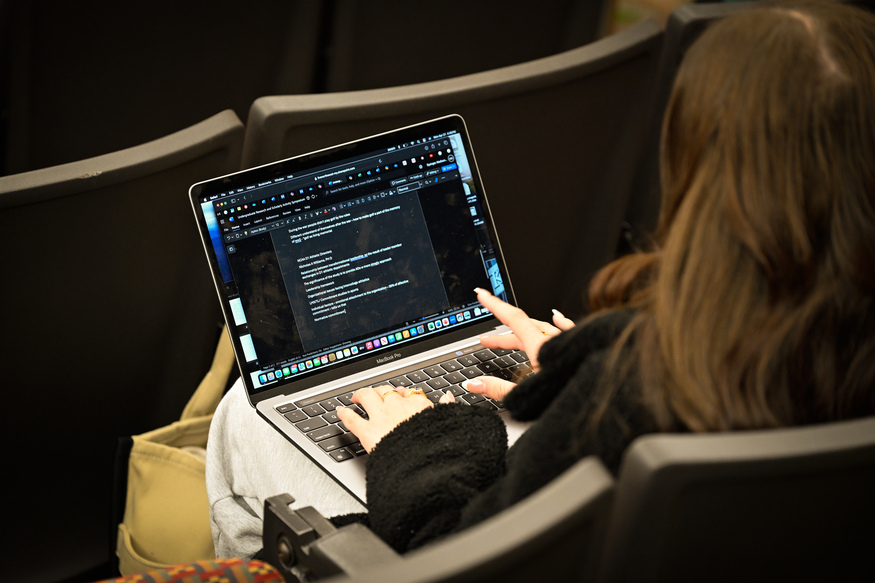 Person sitting in an auditorium seat, using a laptop to take notes on a dark-themed text editor while attending online classes for adults. The screen displays both text and code.