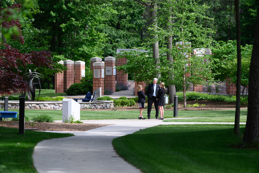 Three people in formal attire stand and talk on a sidewalk in a landscaped, tree-filled campus—perhaps discussing online classes for adults—with monuments and greenery in the background.