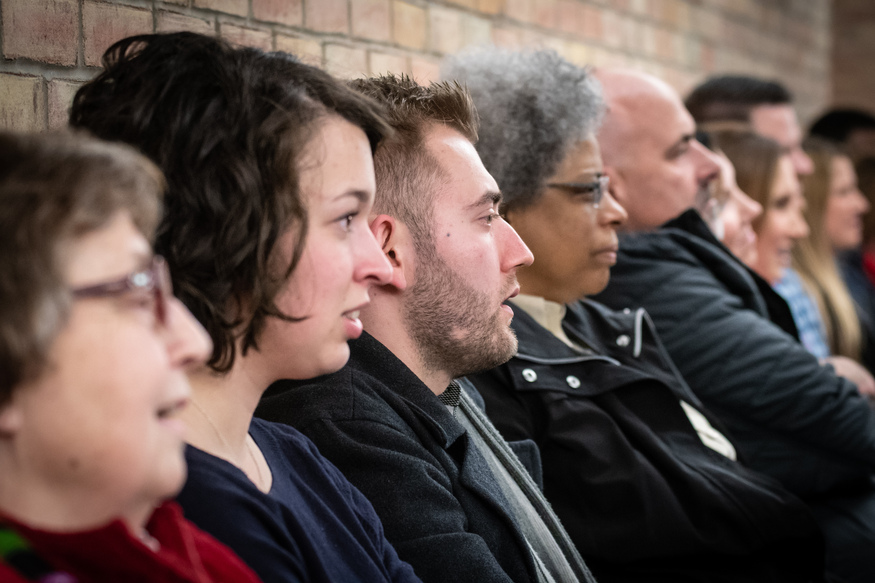 A group of people of various ages sit in a row against a brick wall, facing forward with attentive expressions, as if eagerly anticipating online classes for adults.