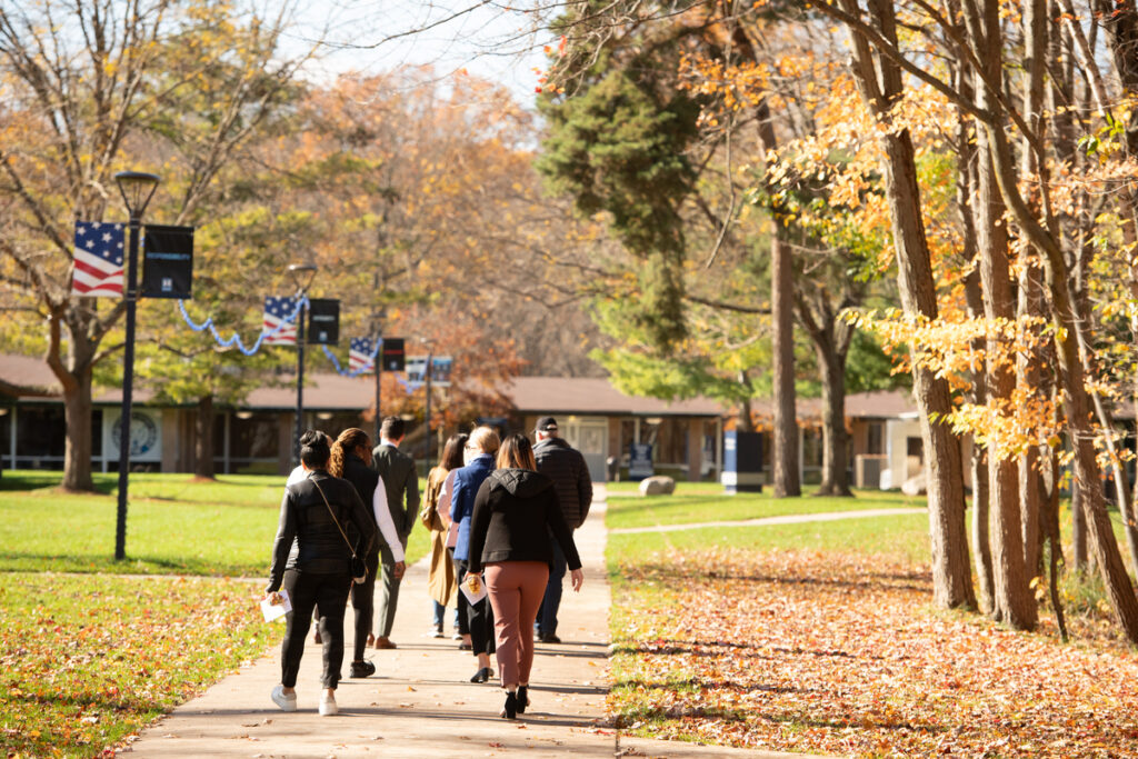 A group of people walk along a tree-lined sidewalk on a sunny autumn day, with fallen leaves on the ground and American flags hanging from lamp posts, discussing online classes for adults.