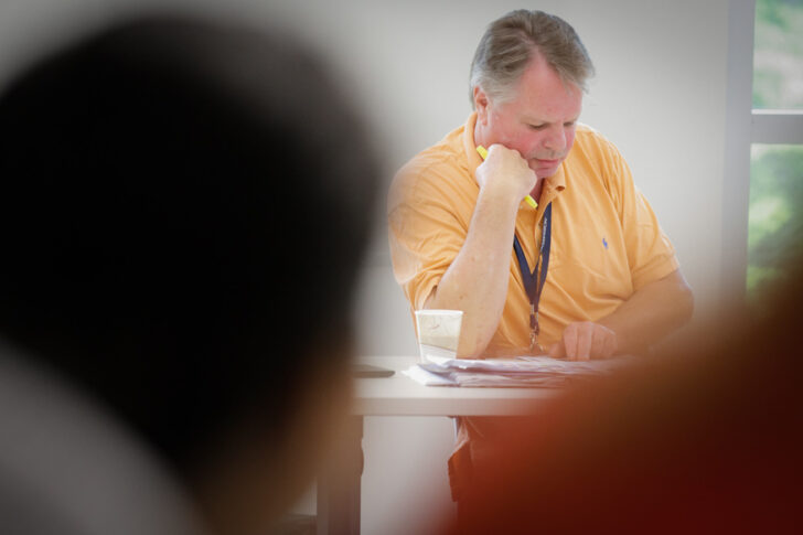 A man in an orange shirt sits at a desk, reading and writing on papers, with a cup and lanyard visible.