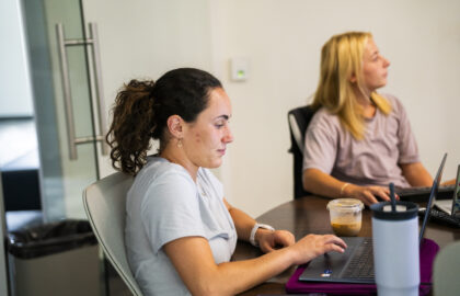Two women sit at a table working on laptops; one is typing, the other looks to the side. A cup of iced coffee and a water bottle are on the table.