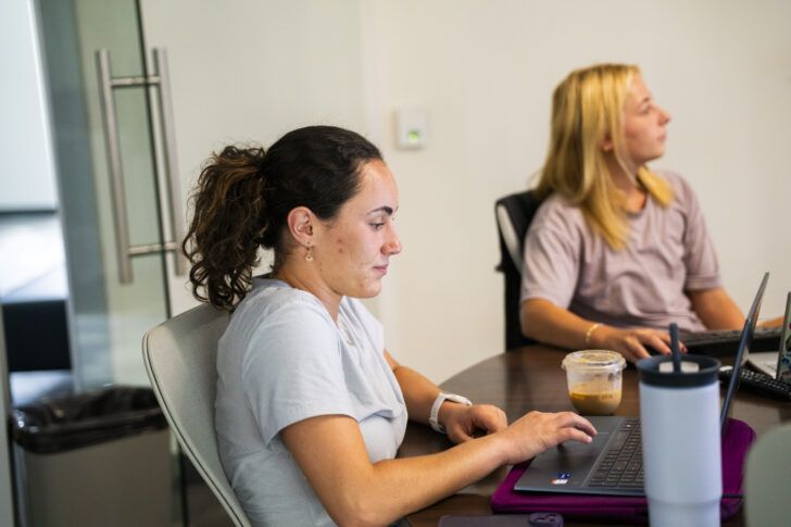 Two women sit at a table working on laptops; one is typing, the other looks to the side. A cup of iced coffee and a water bottle are on the table.