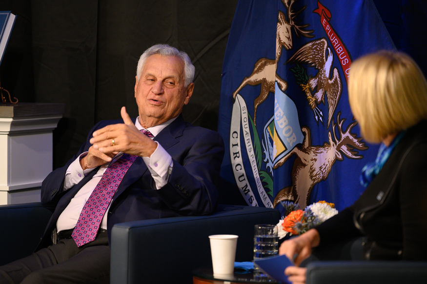 An older man in a suit speaks while seated next to a woman in front of the Michigan state flag, possibly discussing universities in Michigan. A small table with flowers, water, and a cup is visible between them.