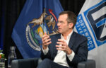 A man in a suit speaks while seated, gesturing with his hands. Michigan state and Northwood University flags are visible in the background.