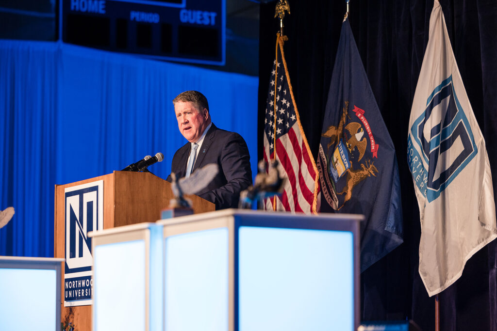 A man in a suit speaks at a podium with Northwood University branding, flanked by the U.S. and Michigan flags at an indoor event highlighting universities in Michigan.