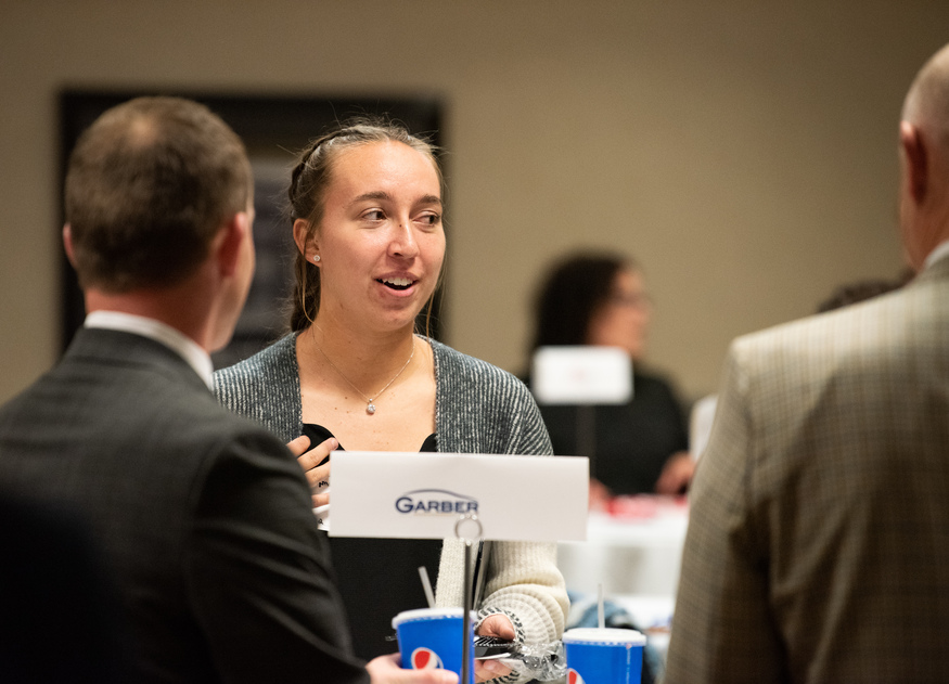 A woman speaks to two men at a networking event, standing near a table with a Garber sign and drinks—an ideal setting for discussing opportunities at universities in Michigan. Other attendees mingle in the background.
