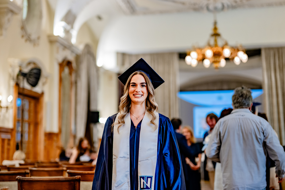 A woman in a blue graduation gown and cap stands smiling in a formal indoor setting, possibly celebrating her achievement at one of the top universities in Michigan, with people and chandeliers in the background.