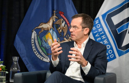 A man in a suit speaks while seated, gesturing with his hands. Michigan state and Northwood University flags are visible in the background.