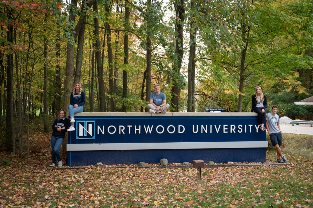 Five people pose on and beside a large "Northwood University" sign in a wooded area with autumn foliage, showcasing one of the vibrant universities in Michigan.