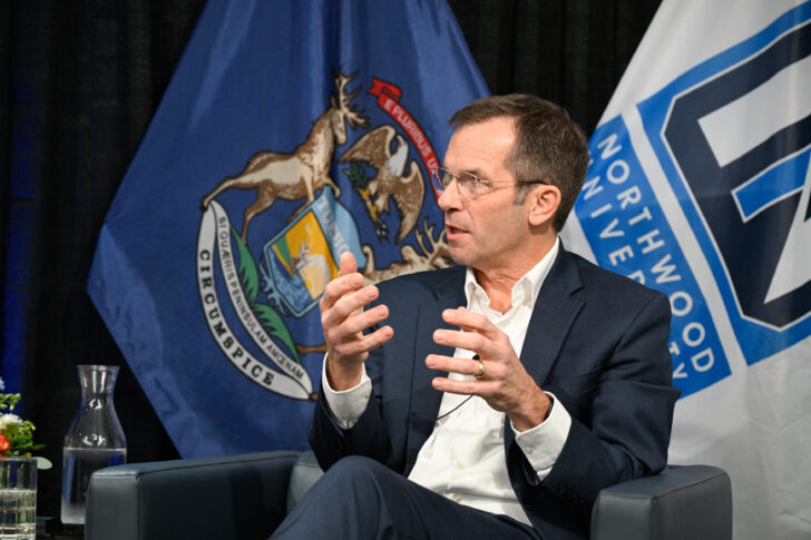 A man in a suit speaks while seated, gesturing with his hands. Michigan state and Northwood University flags are visible in the background.