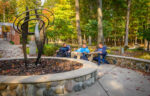 Four people sit and talk on benches near a black metal sculpture surrounded by a stone planter, with trees and autumn foliage in the background.