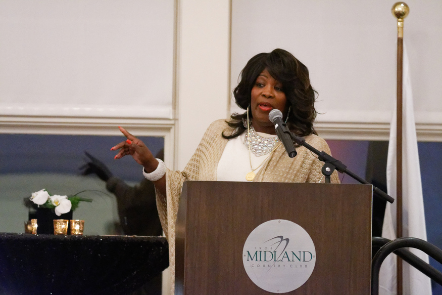 A woman stands at a podium labeled "Midland Country Club," speaking into a microphone and gesturing with her right hand. Beside her, a table holds candles and flowers, as she addresses attendees from a university in Michigan, USA.