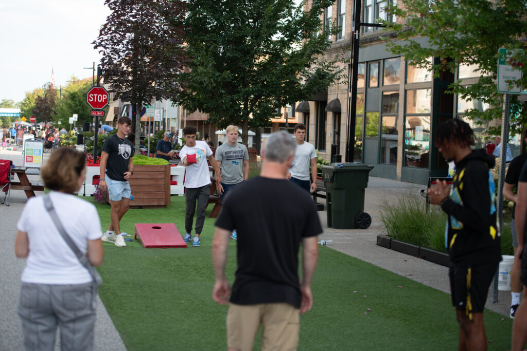 A group of people stands and plays cornhole on a city street lined with buildings near a university in Michigan, USA; some watch while others walk by.