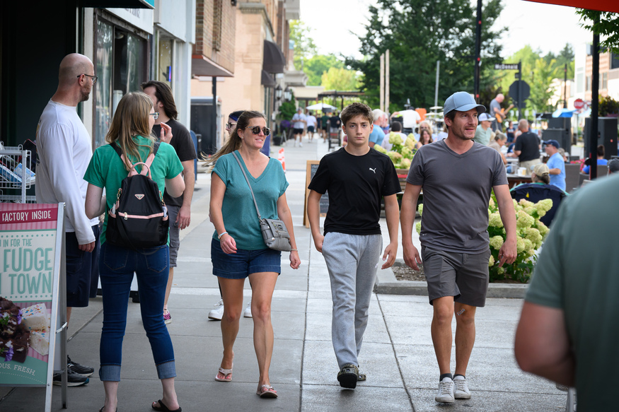 People walk along a busy sidewalk in a downtown area near a university in Michigan, USA, enjoying the sunny day as they pass storefronts and outdoor seating.