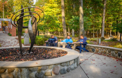 Four people sit and talk on benches near a black metal sculpture surrounded by a stone planter, with trees and autumn foliage in the background.