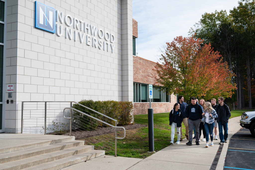 A group of students walk on a sidewalk outside a building with a "Northwood University" sign on the wall.
