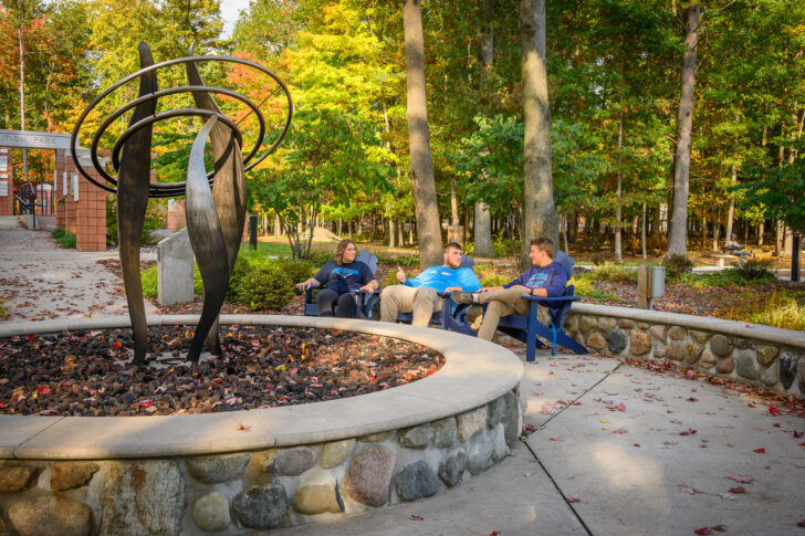 Four people sit and talk on benches near a black metal sculpture surrounded by a stone planter, with trees and autumn foliage in the background.