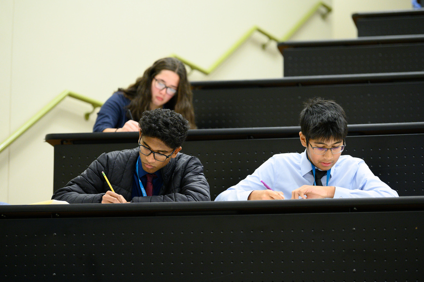Three students sit in a lecture hall at one of the top undergraduate business schools, spaced apart, focused on writing or taking a test at their desks.