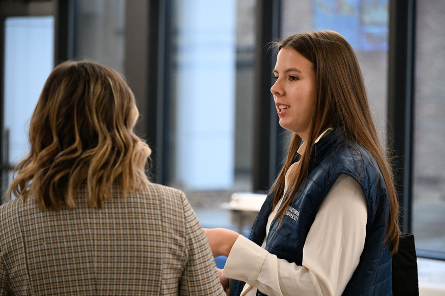 Two women are having a conversation indoors; one, possibly discussing top undergraduate business schools, is facing the camera in a blue vest, while the other, in a plaid jacket, has her back to the camera.