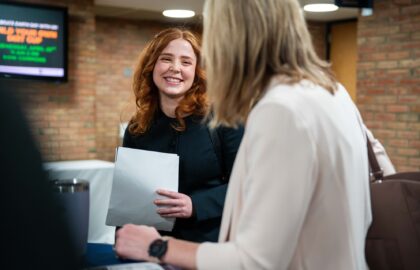 A woman with red hair holds a folder and smiles while talking to another person in an indoor setting.