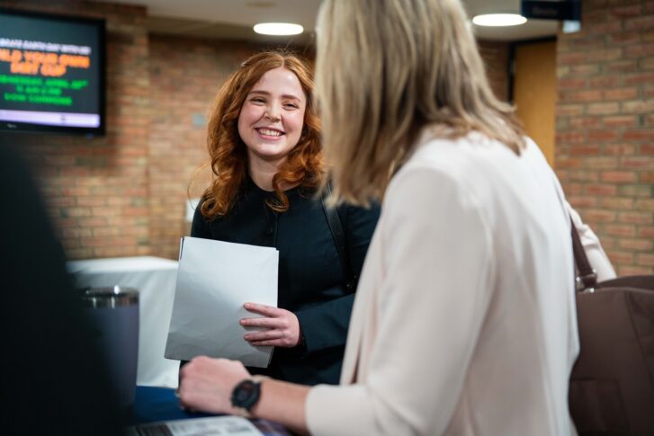 A woman with red hair holds a folder and smiles while talking to another person in an indoor setting.