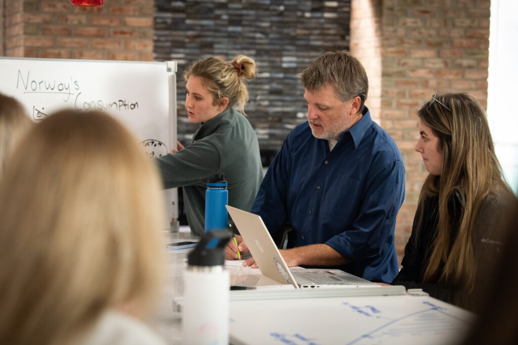Three people sit at a table with laptops, while one person writes on a whiteboard labeled "Norway's Consumption" during a group discussion.